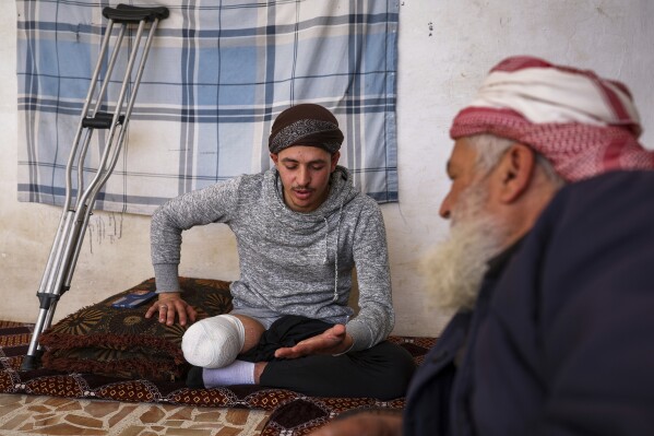 Suleiman Khalil, 21, who lost his leg in a landmine explosion while harvesting olives with his friends in a field, talks with his father at their home in the village of Qaminas, east of Idlib, Syria, Wednesday, April 9, 2025. (AP Photo/Ghaith Alsayed)