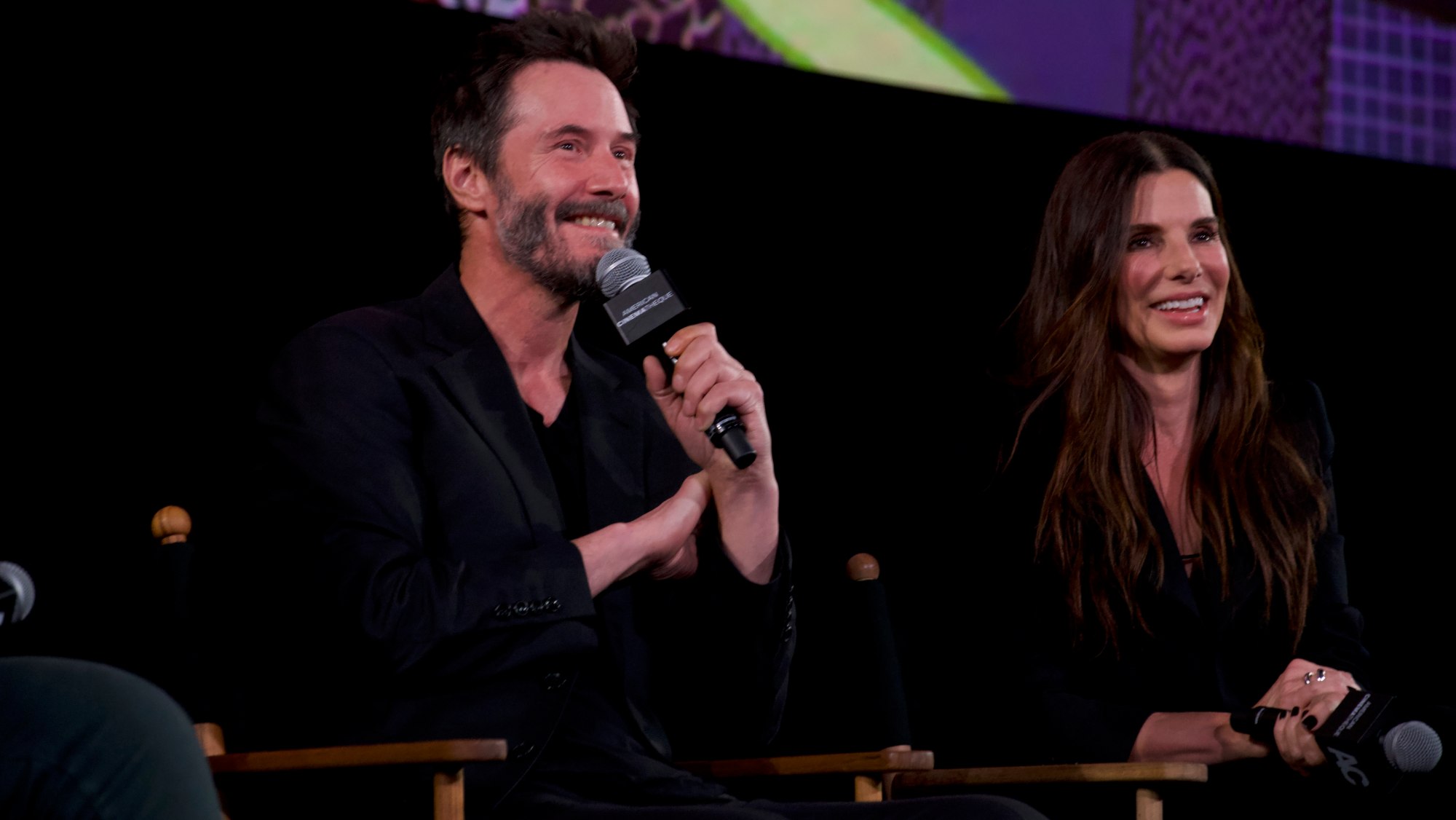 Keanu Reeves holds a microphone while Sandra Bullock smiles next to him